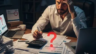 Um homem com a mão na cabeça, sentado ao redor de uma mesa a sua frente um calculadora e um ponto de exclamação sinalizando preocupação com as contas